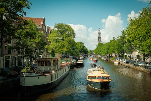 Scenic view of Amsterdam's iconic canal with historic boats and lush greenery under a bright blue sky.