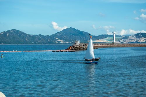 Free A peaceful scene of a sailboat on calm waters with a lighthouse and mountains in the background. Stock Photo