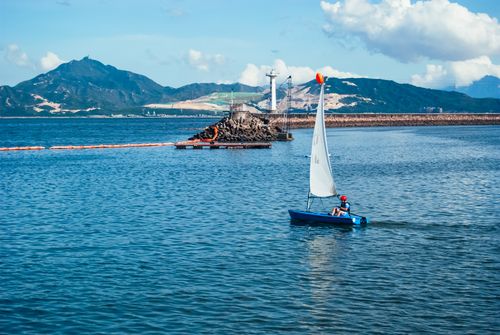 Free A lone sailboat navigates near a coastal lighthouse with scenic mountain backdrop. Stock Photo