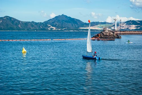 Free A person sailing on a small boat on calm waters with mountains and a lighthouse in the background. Stock Photo