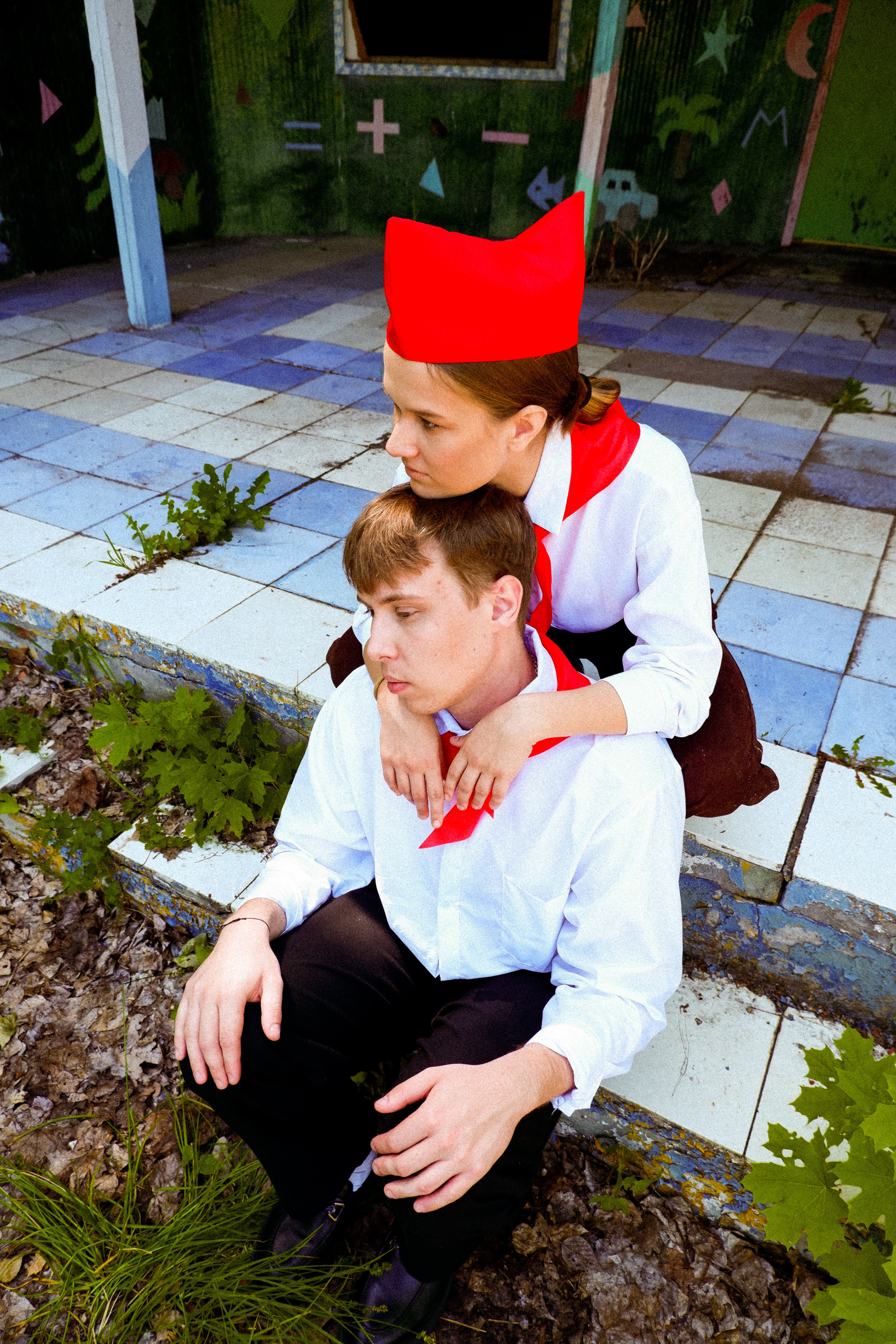 Young couple in vintage uniforms sitting in an overgrown, abandoned tiled outdoor area.