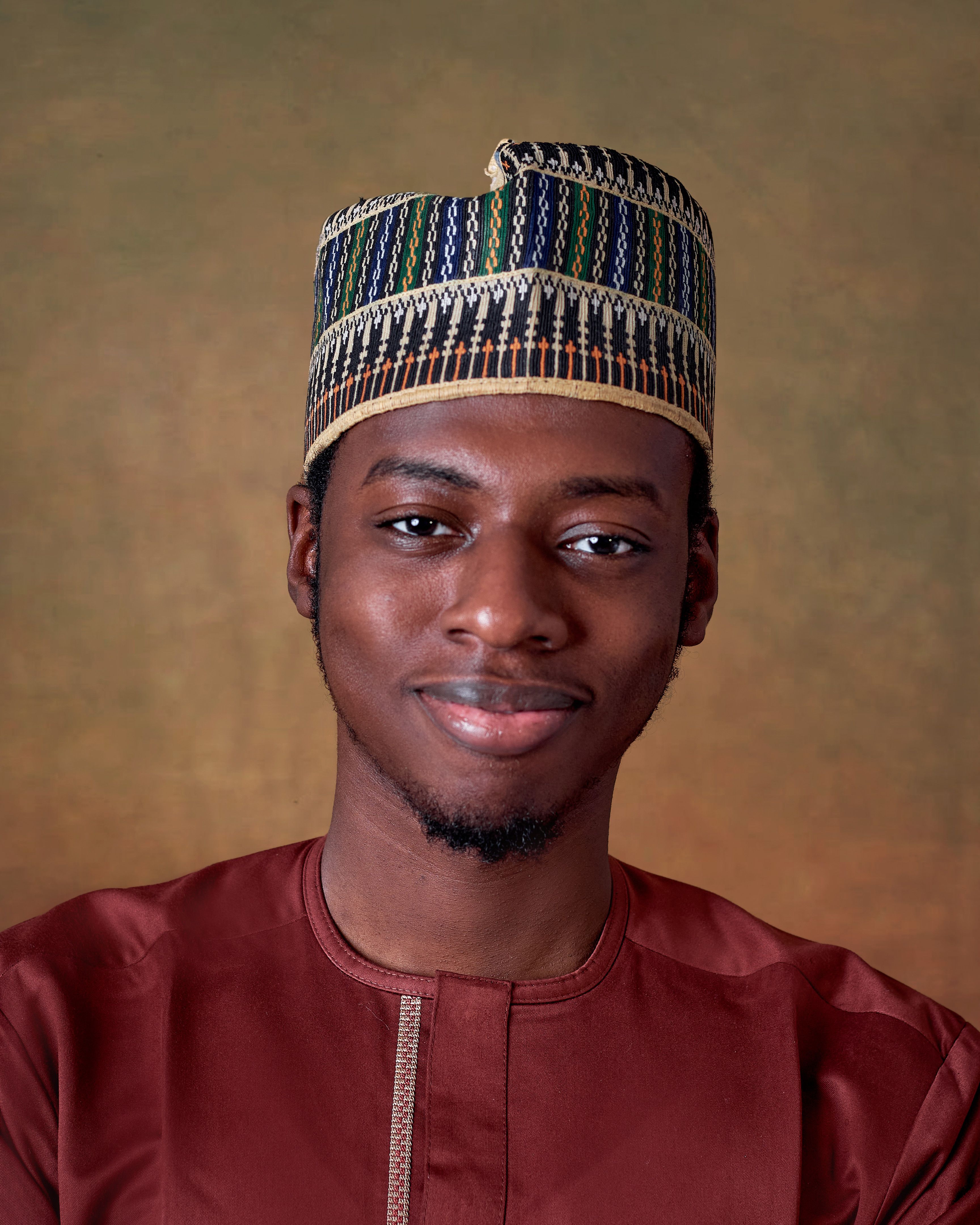 Smiling young man wearing traditional African attire and cap on a neutral background.