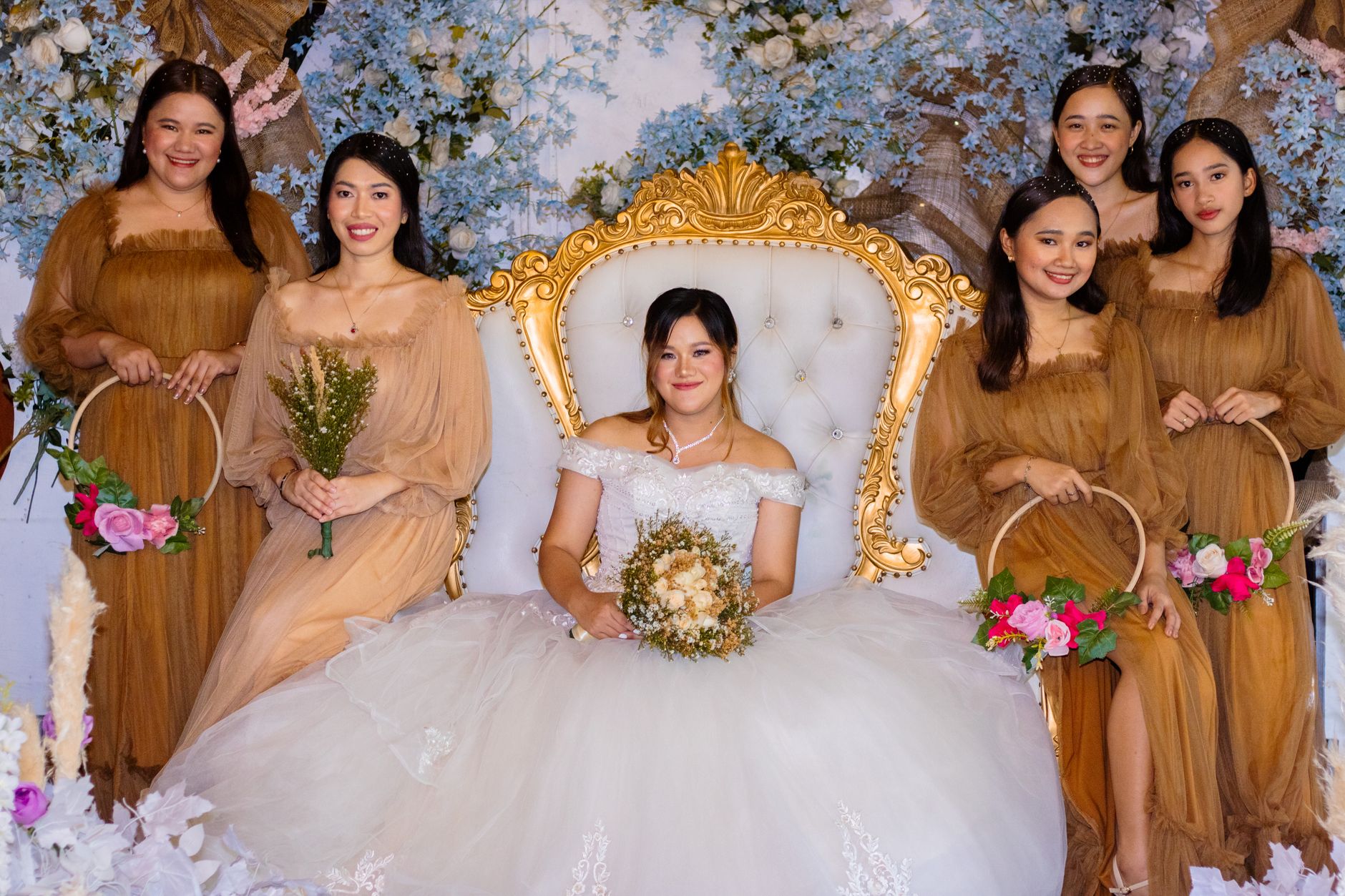 A bride and her bridesmaids pose in elegant dresses against a floral backdrop, creating a perfect wedding moment.