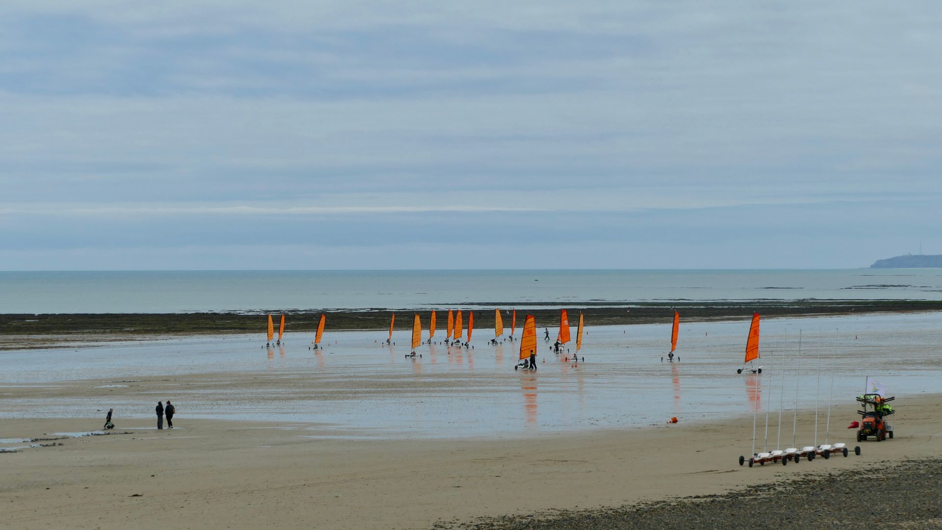 People enjoying beach sailing at Port-Bail-sur-Mer, France with vibrant orange sails reflecting on wet sand.