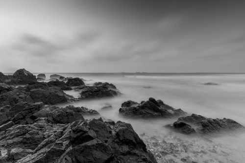 Free Serene black and white photograph of a rocky shoreline with smooth ocean waves at twilight. Stock Photo