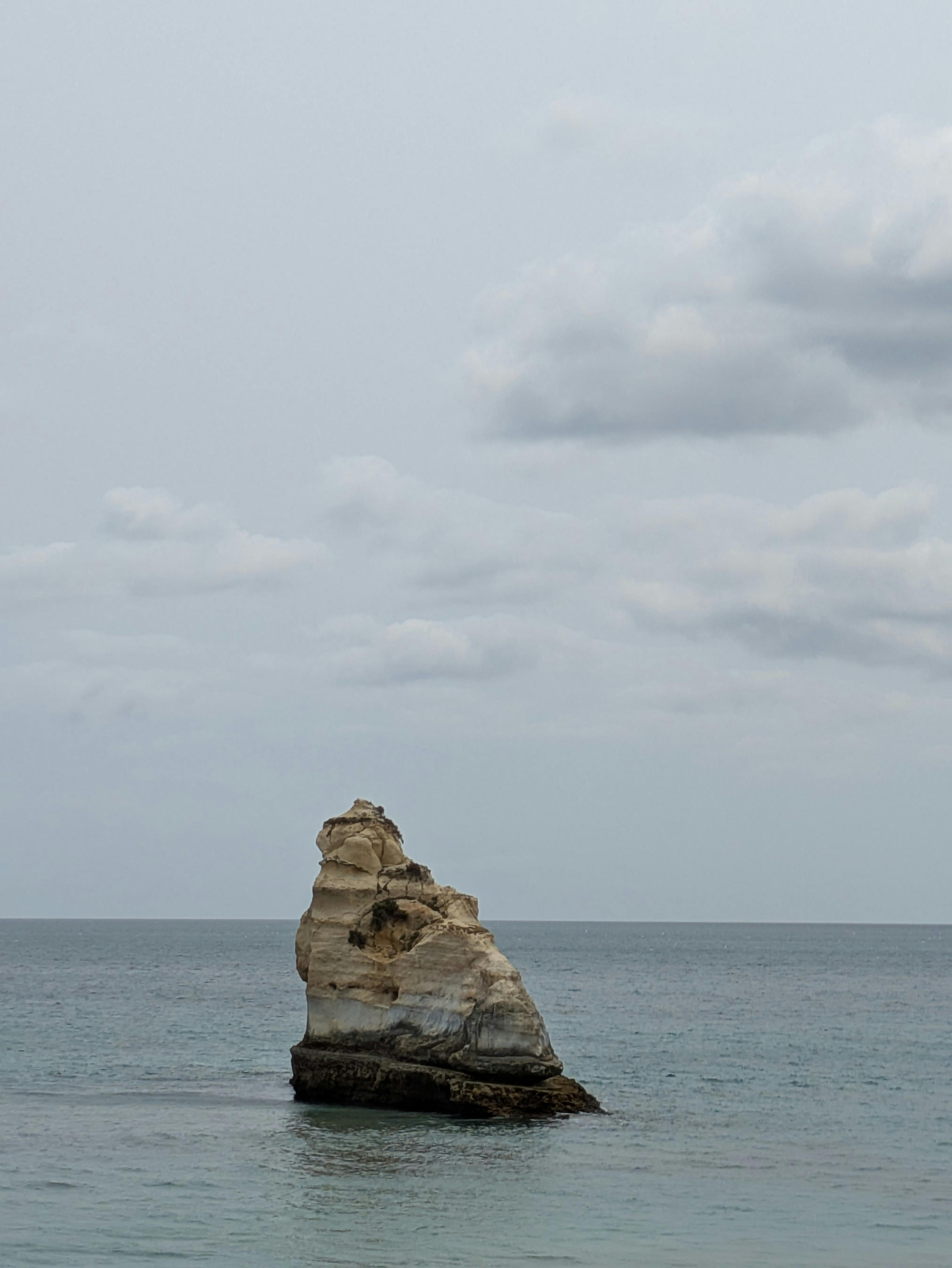 Eroded rock formation in Faro District, Portugal, stands alone in calm ocean under overcast sky.