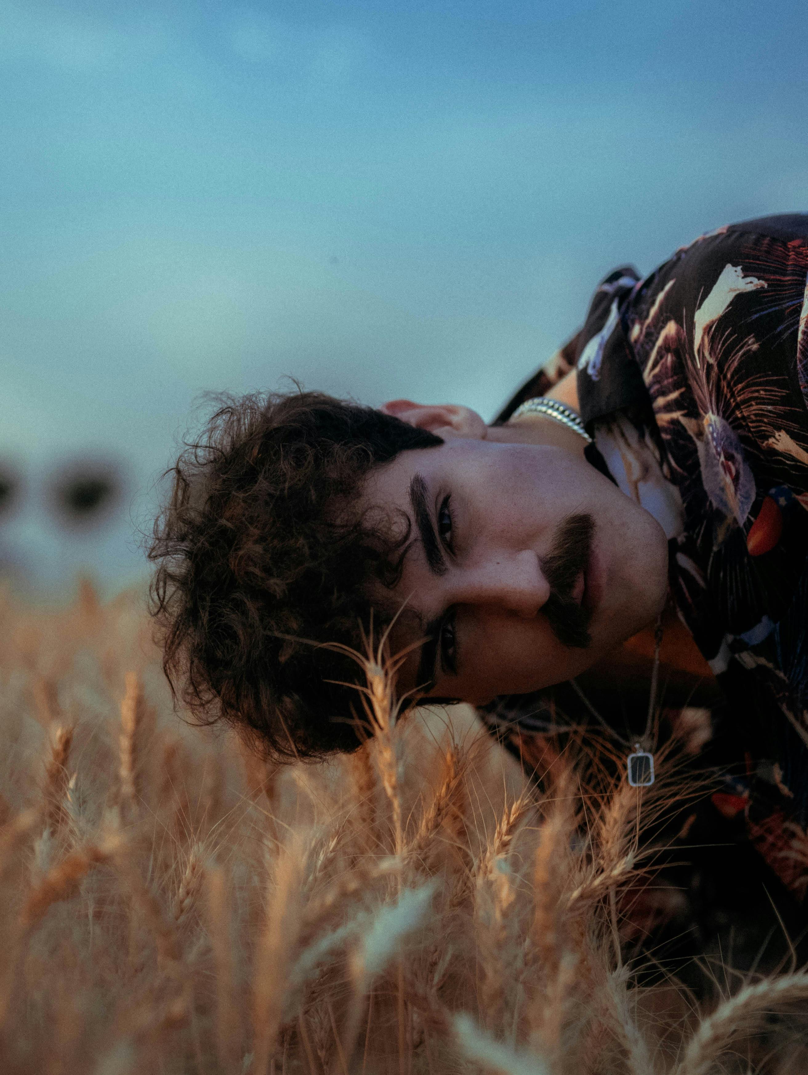 A young man in a floral shirt lies in a wheat field during twilight, creating a serene and pensive atmosphere.