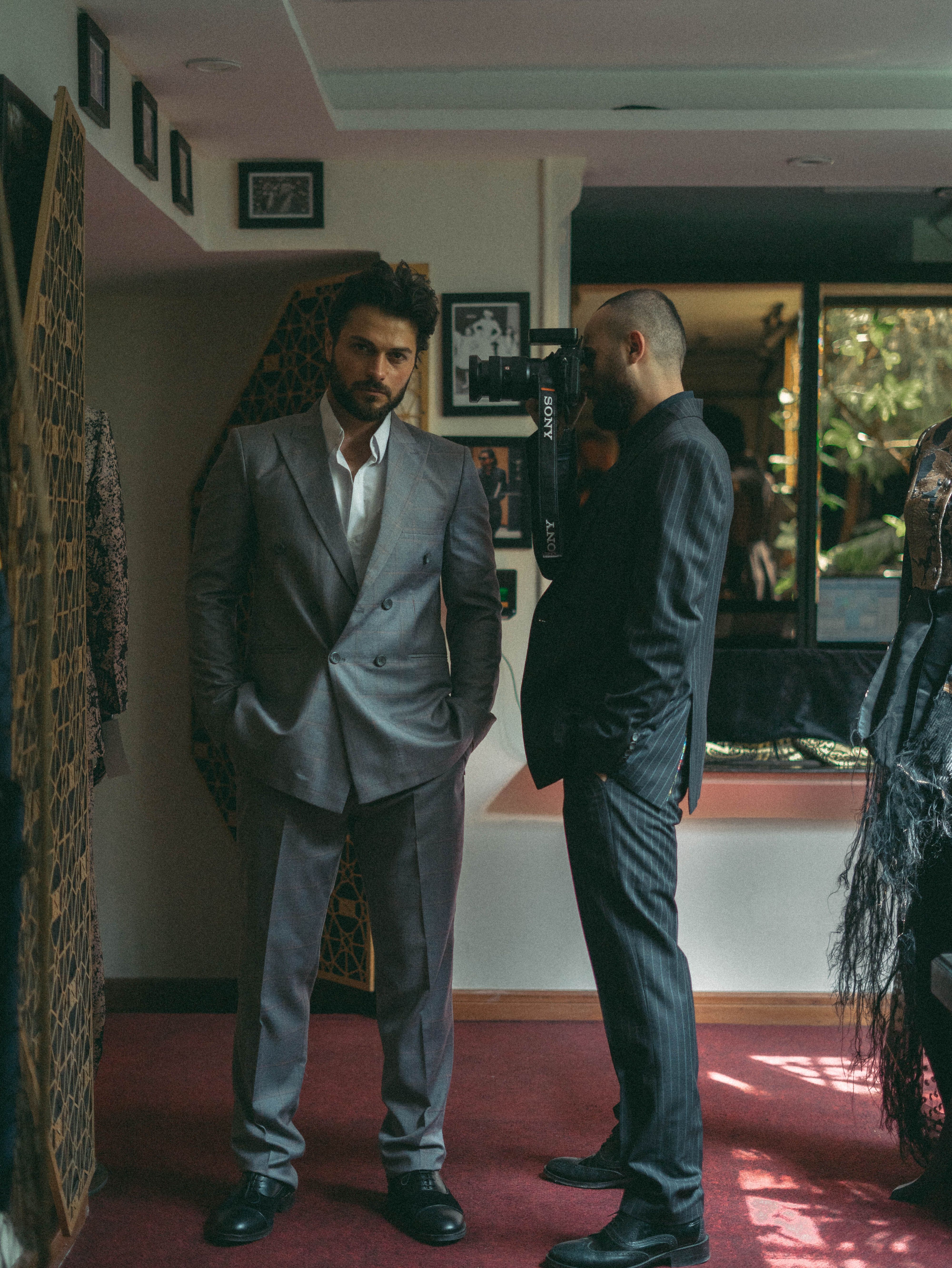 Two men in stylish suits pose in a sophisticated indoor setting. Moody and elegant ambiance.