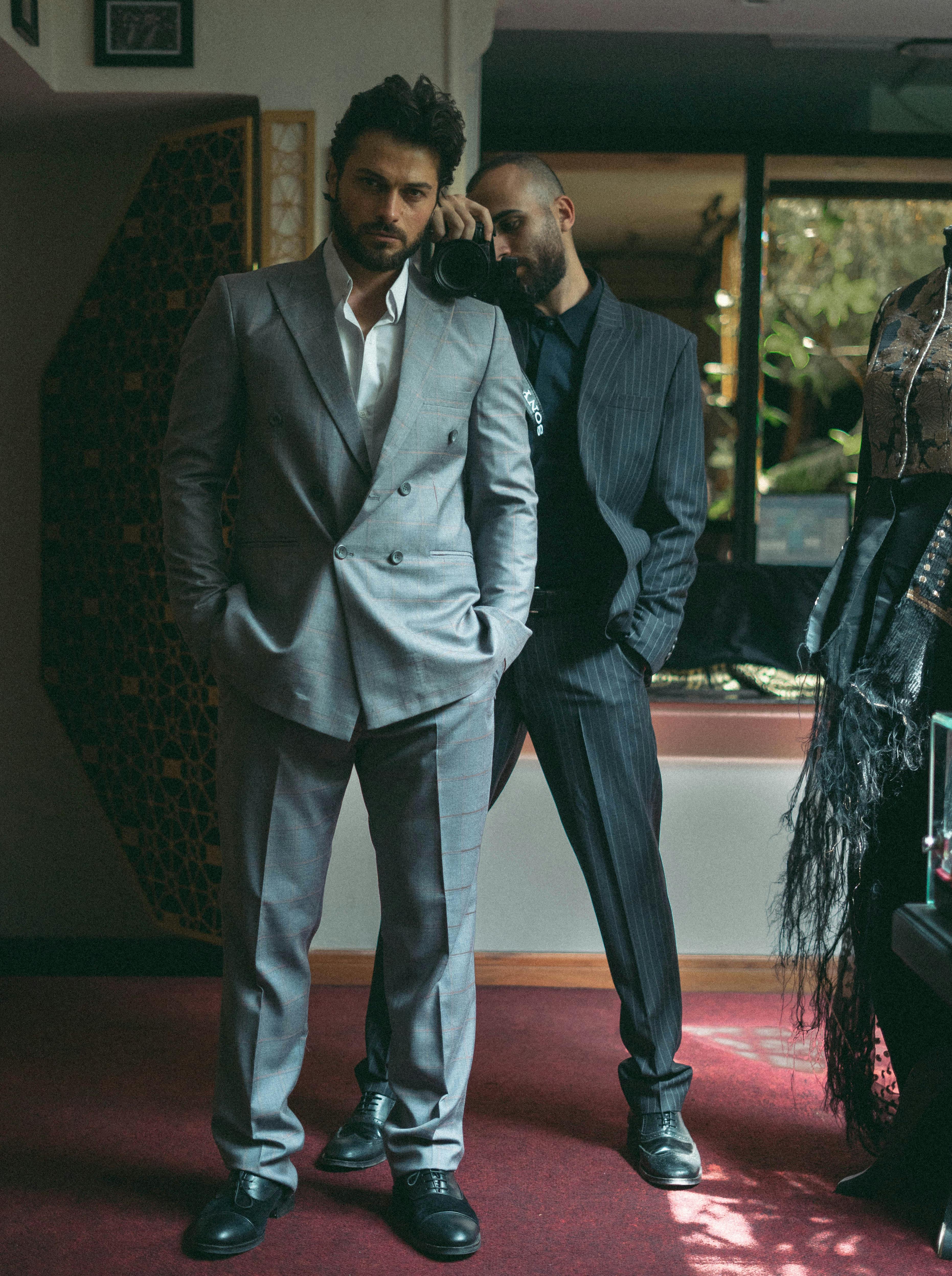 Two men in elegant suits pose indoors, showcasing modern fashion.