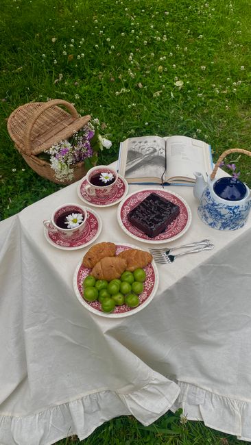 Elegant outdoor picnic setup with tea, croissants, fresh fruit, and flowers on a grassy backdrop.