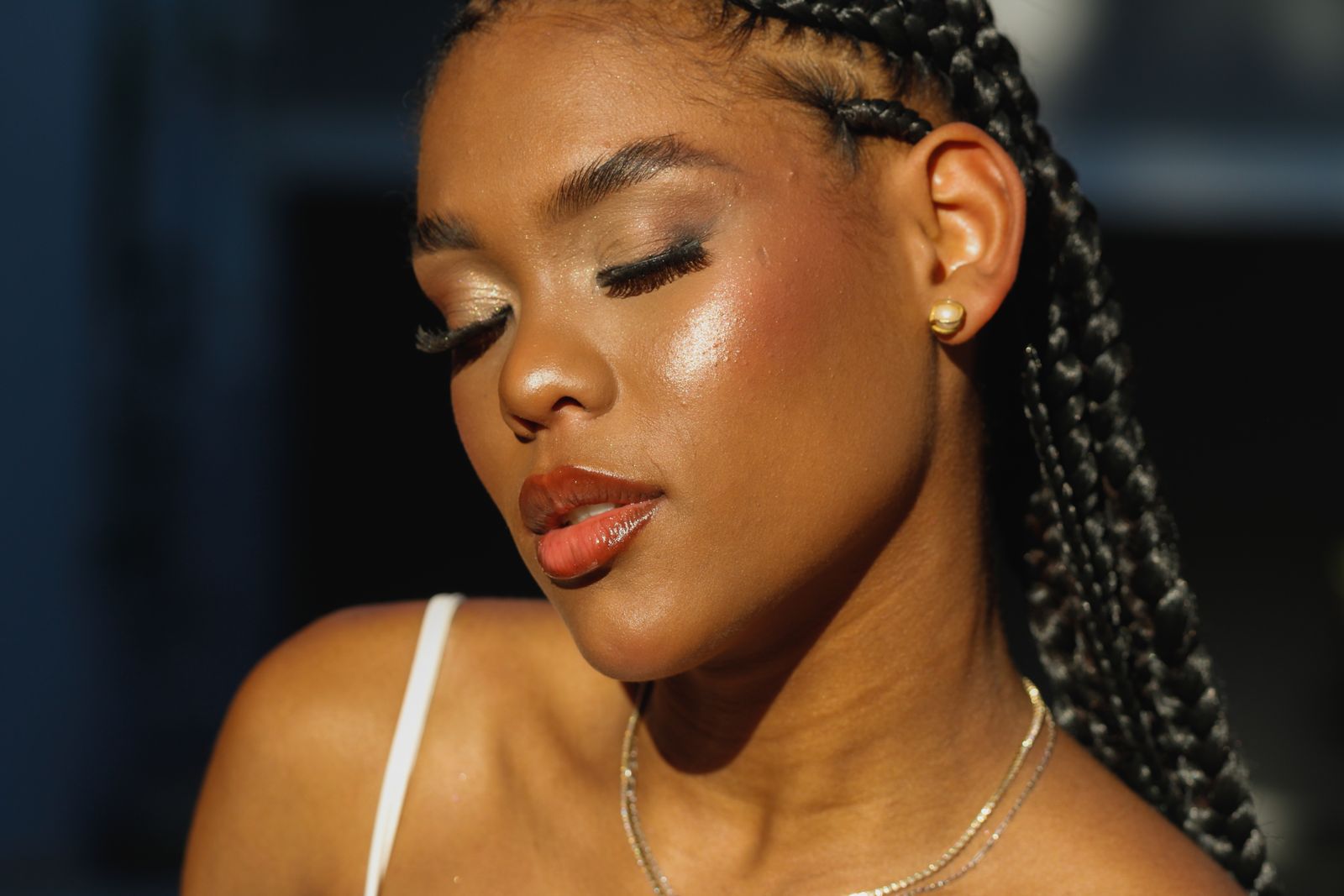 Woman with long braids and closed eyes, glowing skin lit by warm sunlight, wearing gold necklaces