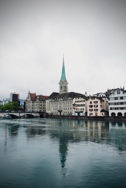 Scenic view of Zurich city skyline with Fraumünster Church reflecting on water under cloudy sky.