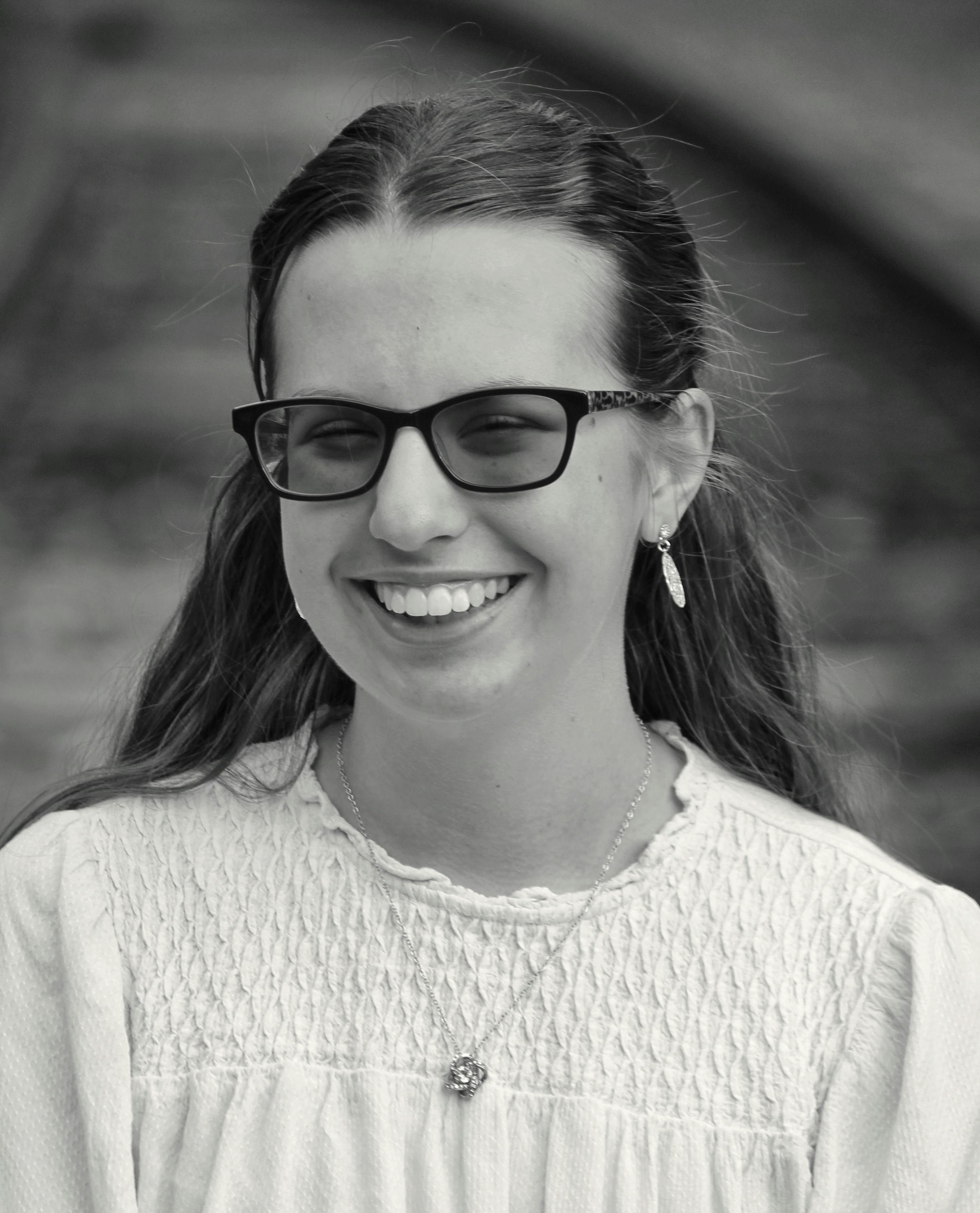 A cheerful young woman wearing glasses in a black and white portrait outdoors.