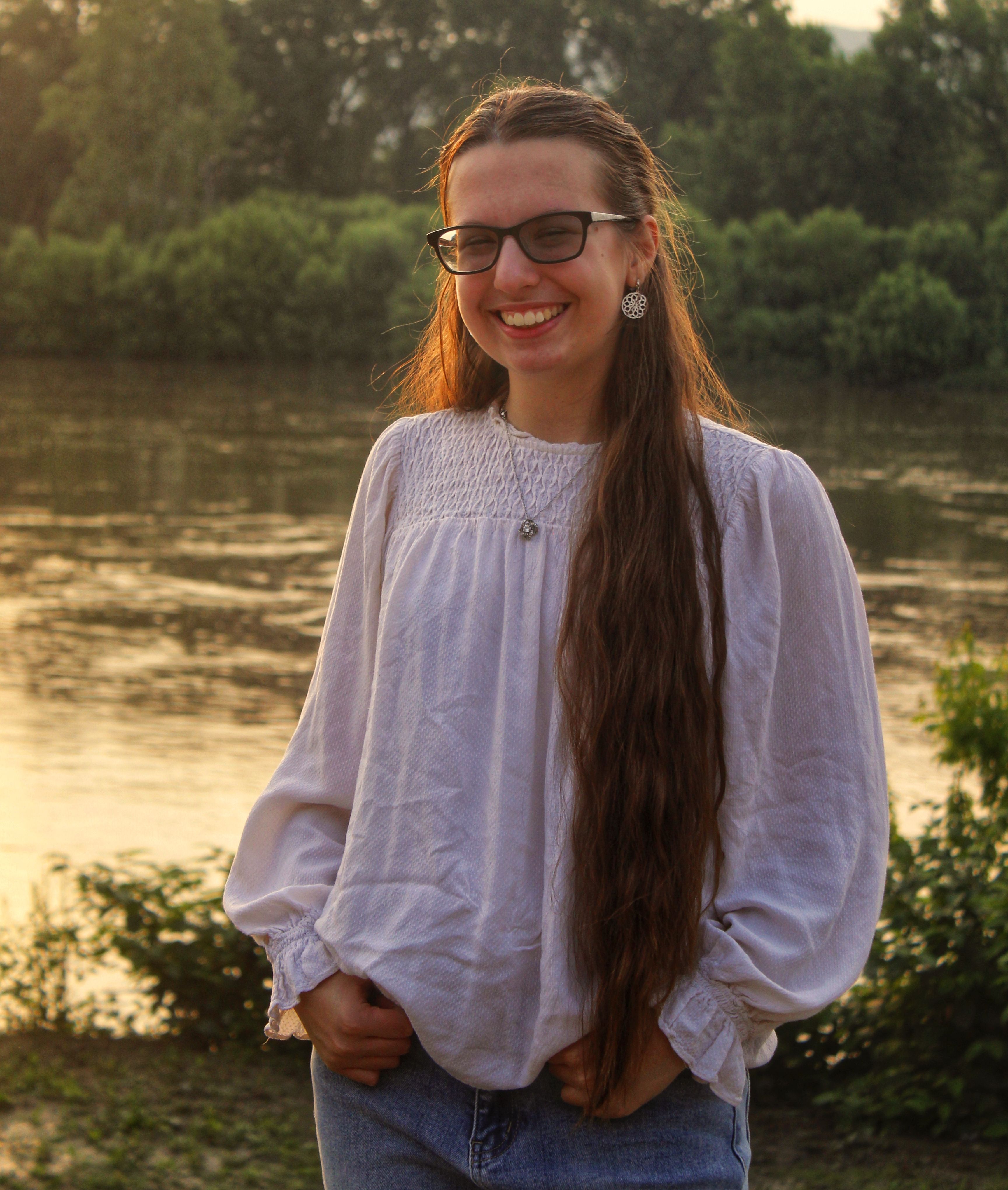 A young woman with long hair smiling by a river at sunset, wearing a white blouse and glasses.