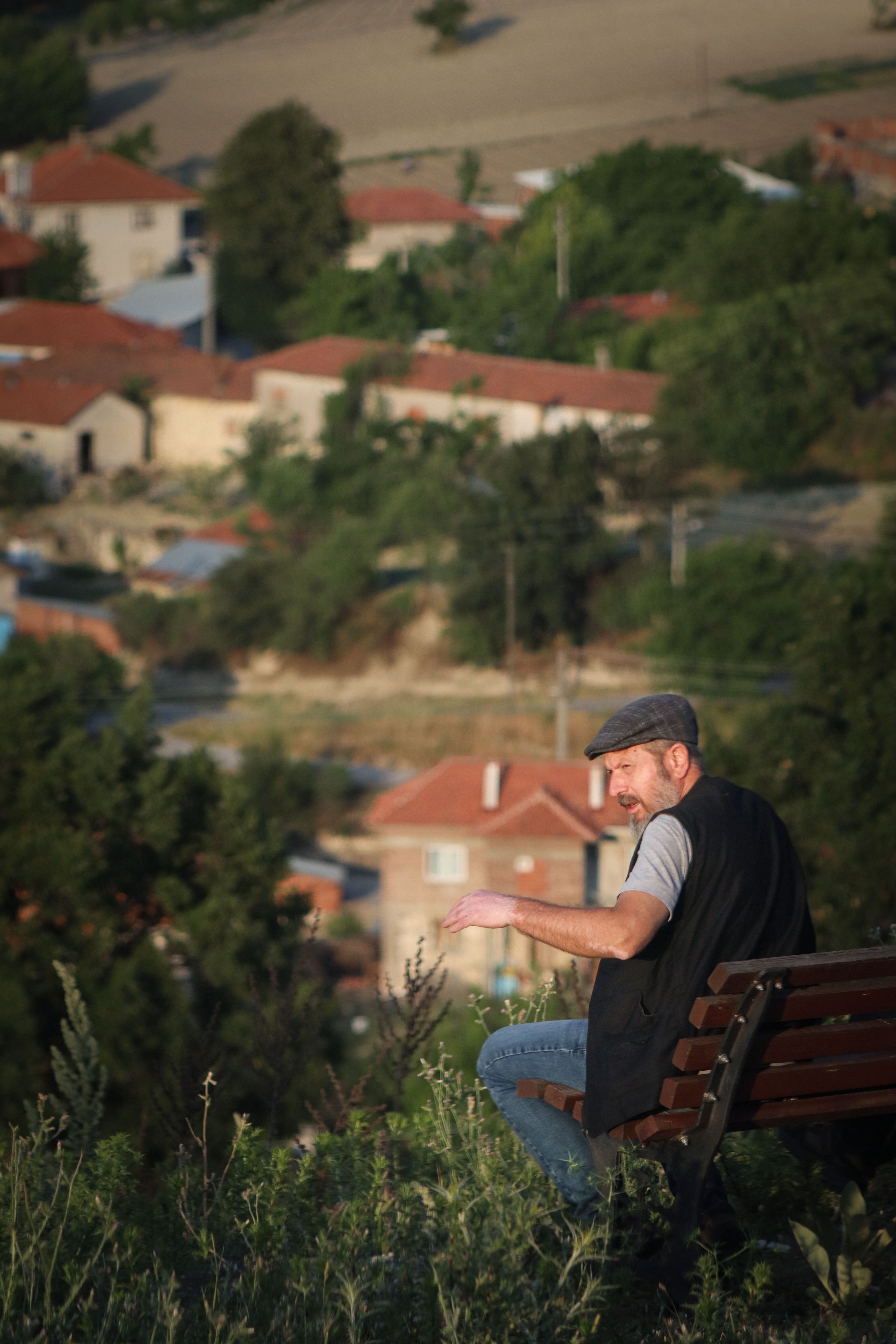 Man seated on a bench enjoying a scenic view of a rural village under sunlight.