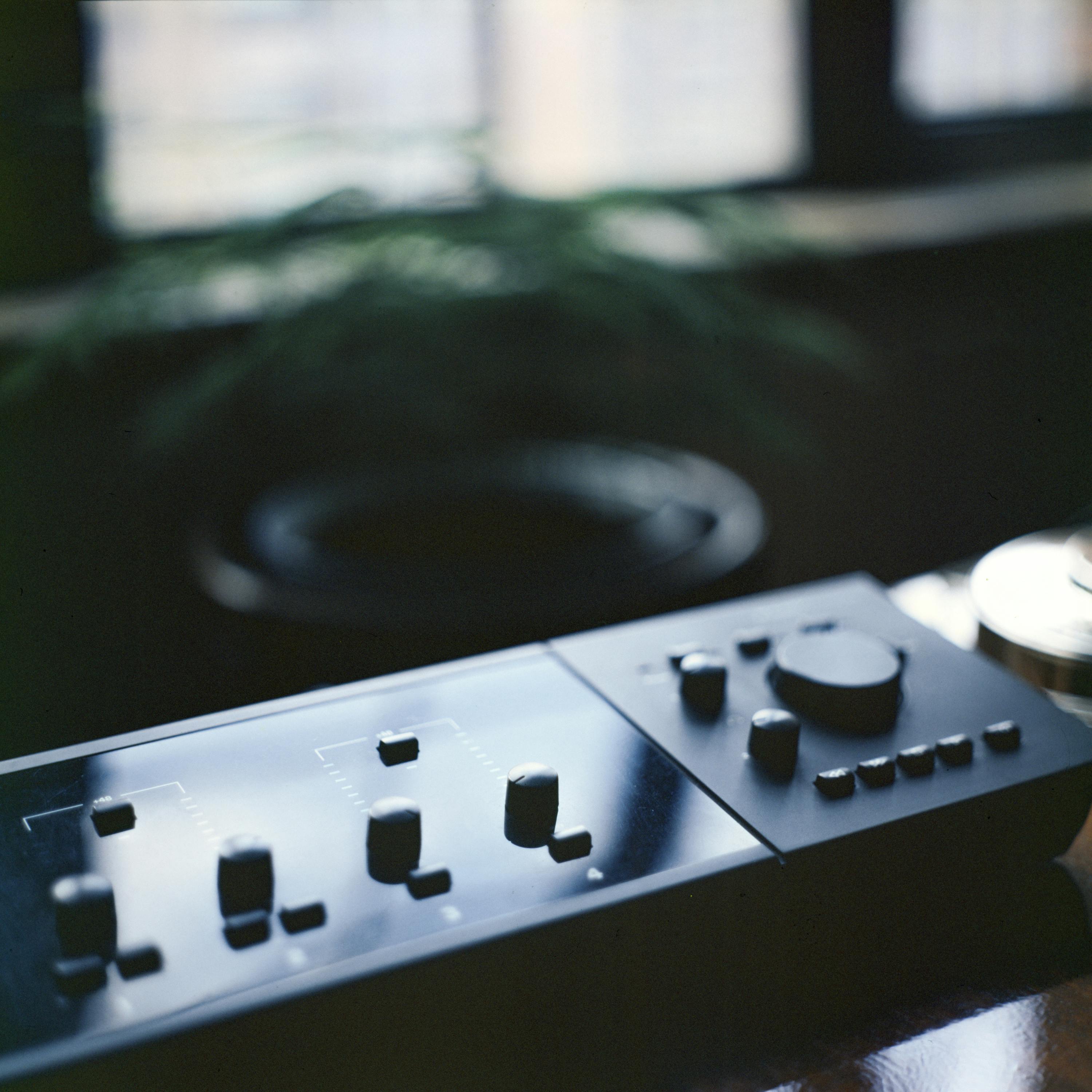 A sleek audio mixer placed on a desk in a softly lit room, with a blurred plant in background.