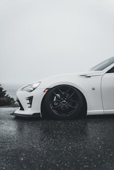 Side view of a stylish white sports car with dark wheels parked on a wet road on a cloudy day.