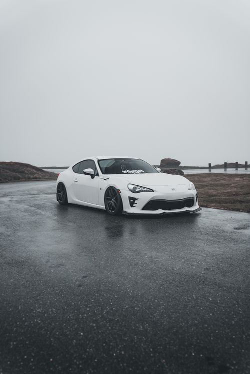 Free A white sports car parked on wet pavement, surrounded by misty weather and overcast skies. Stock Photo