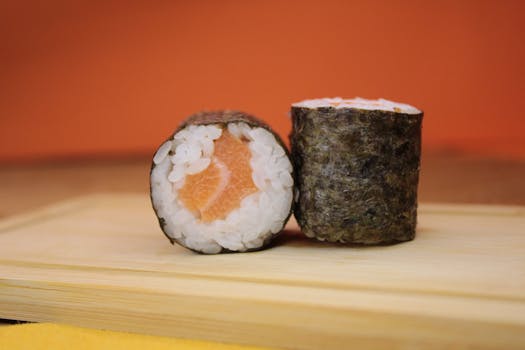 A vibrant close-up of two salmon sushi rolls on a wooden plate against an orange background.