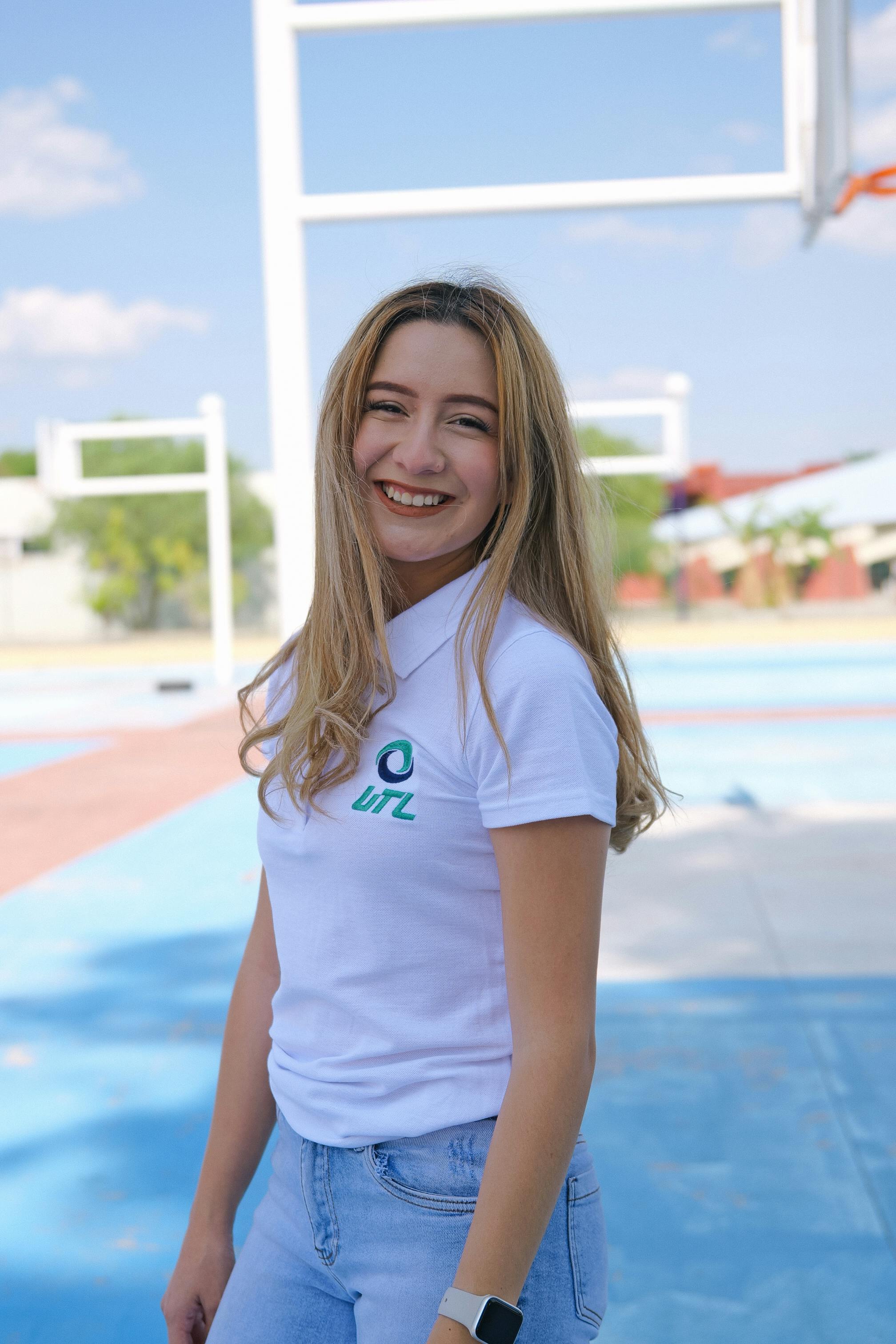 A cheerful young woman smiles outdoors on a sunny day, wearing a white t-shirt and jeans.