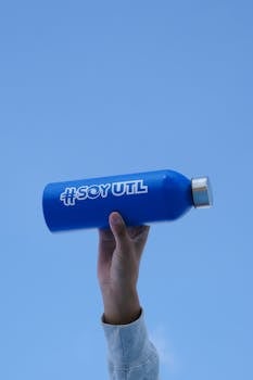 A hand holding a blue water bottle labeled #SoyUTL against a clear sky in León, Mexico.