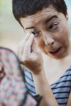Woman applying makeup while looking into a compact mirror outdoors.