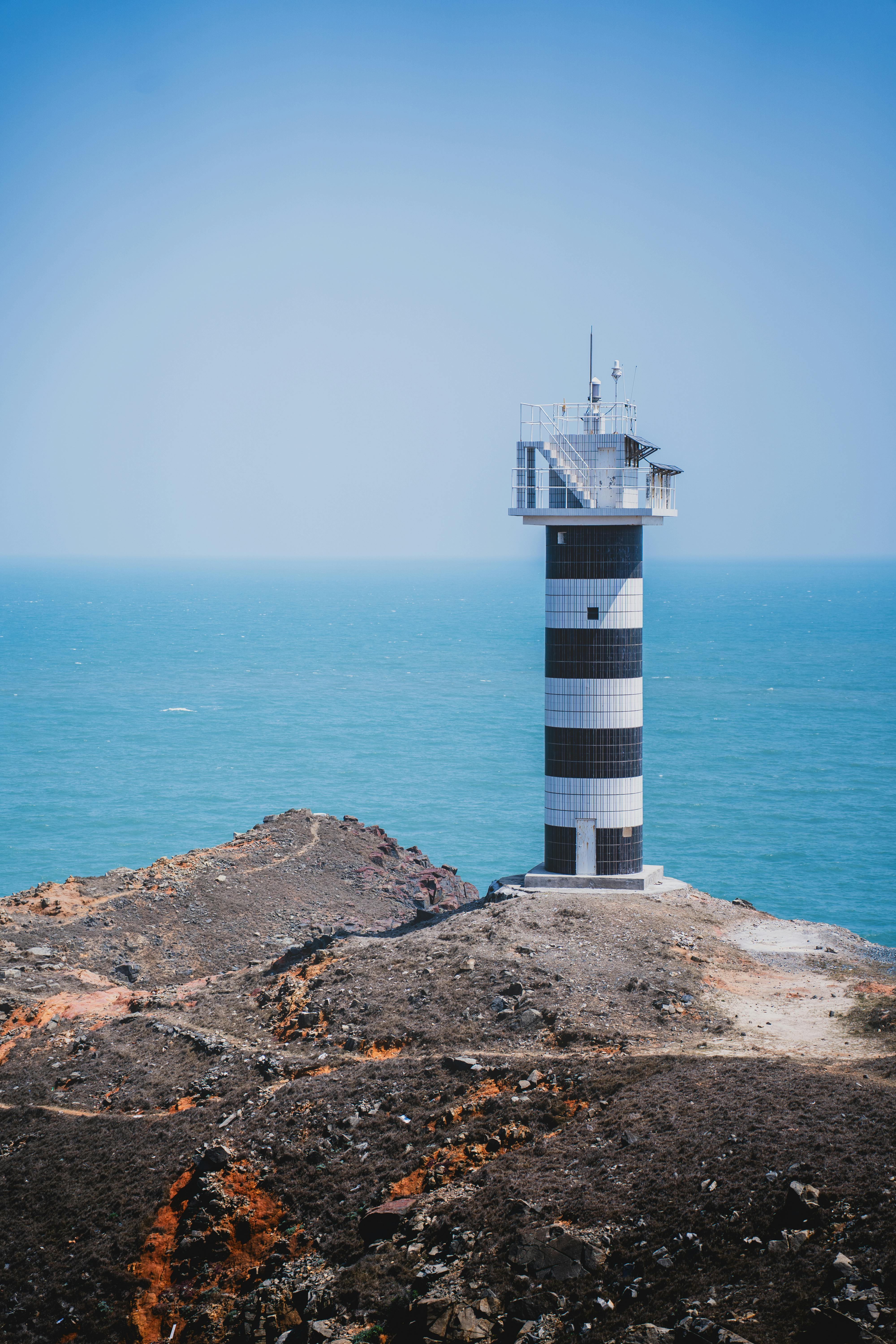 A distinct black and white striped lighthouse overlooking the blue ocean on a rocky coastline.