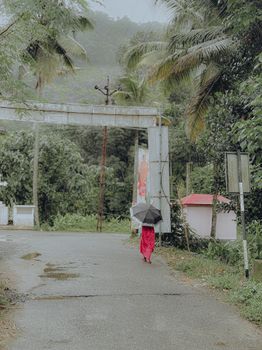 A woman in a vibrant dress walks down a rainy street in Mamalakandam, Kerala, India, with an umbrella.