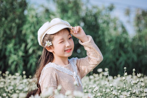 Selective Focus Photography of Girl Wearing Cap Standing Beside Flowers
