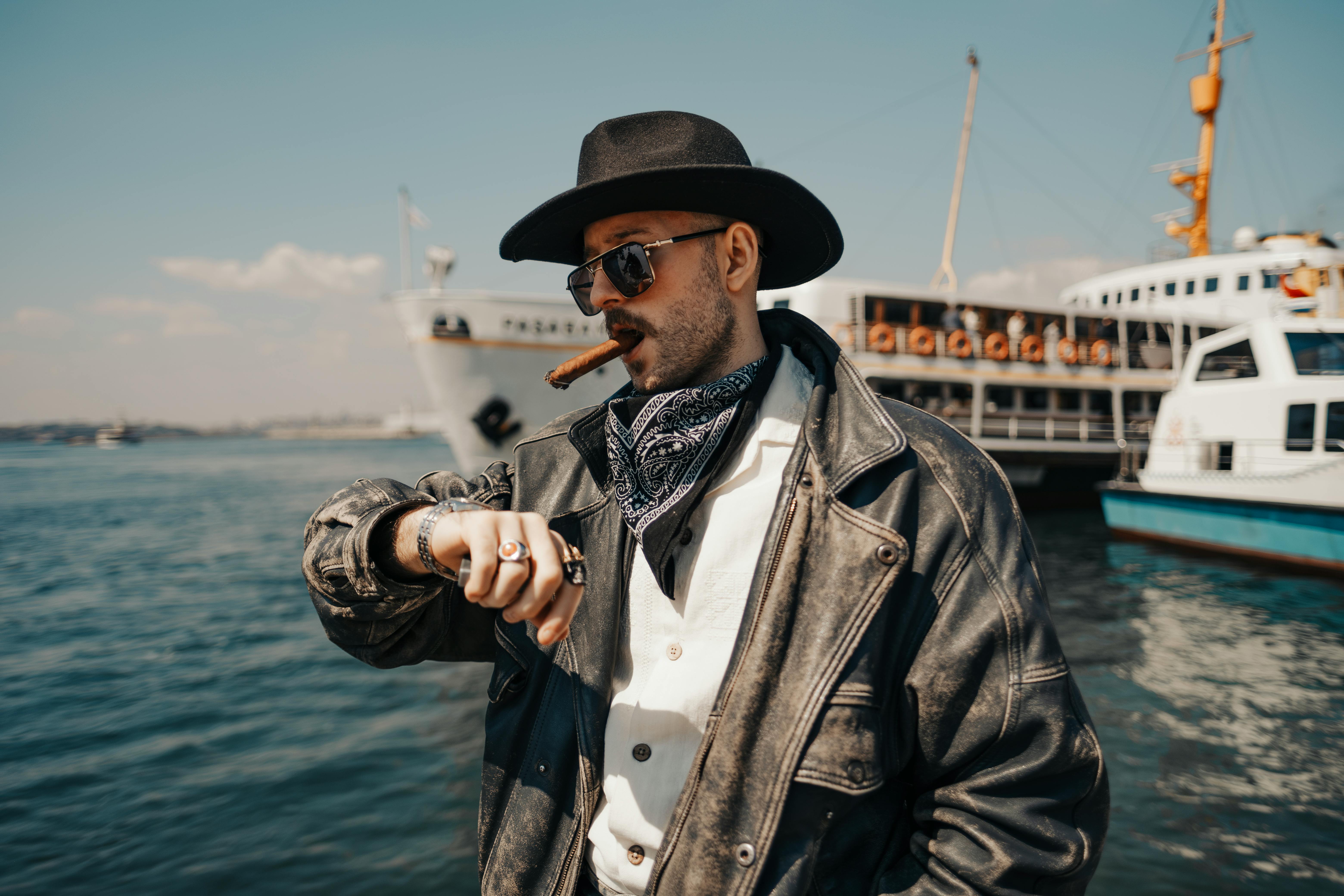Confident man in leather jacket, hat, and sunglasses by the harbor, enjoying a cigar.