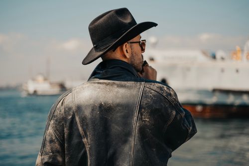 Free A stylish man in a black hat and leather jacket, enjoying a view by the sea with ships in the background. Stock Photo
