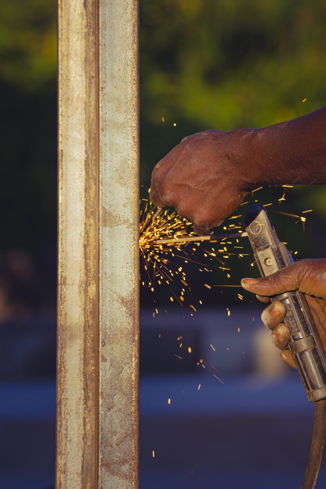 Two hands running a cutting torch along a rusted steel beam outdoors, throwing a dense shower of orange sparks