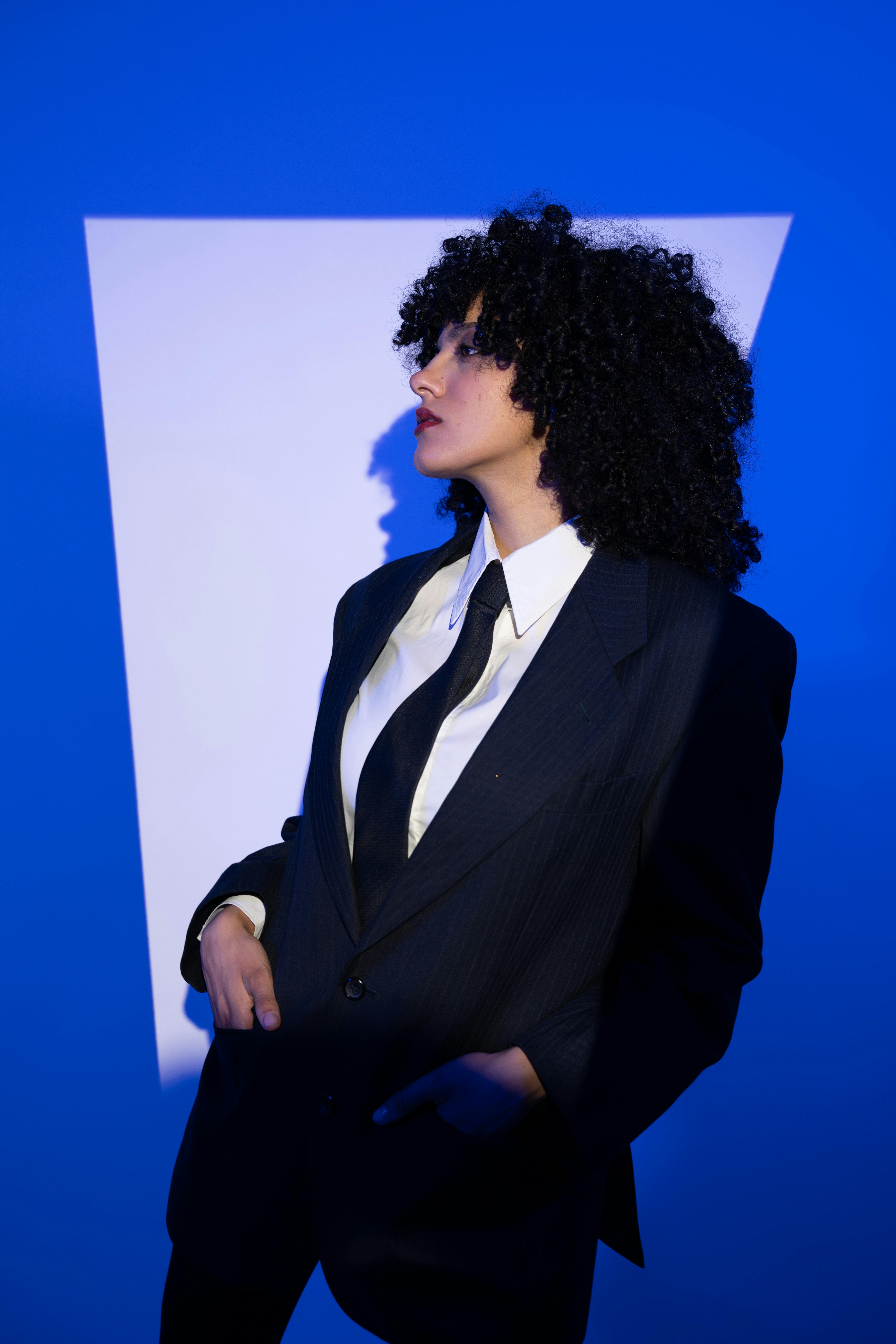 Portrait of a woman with curly hair in a suit, lit against a geometric blue backdrop.