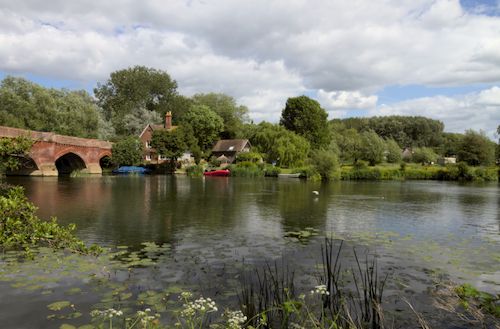 Free A tranquil scene of a river with a red brick bridge and lush greenery under a blue sky. Stock Photo