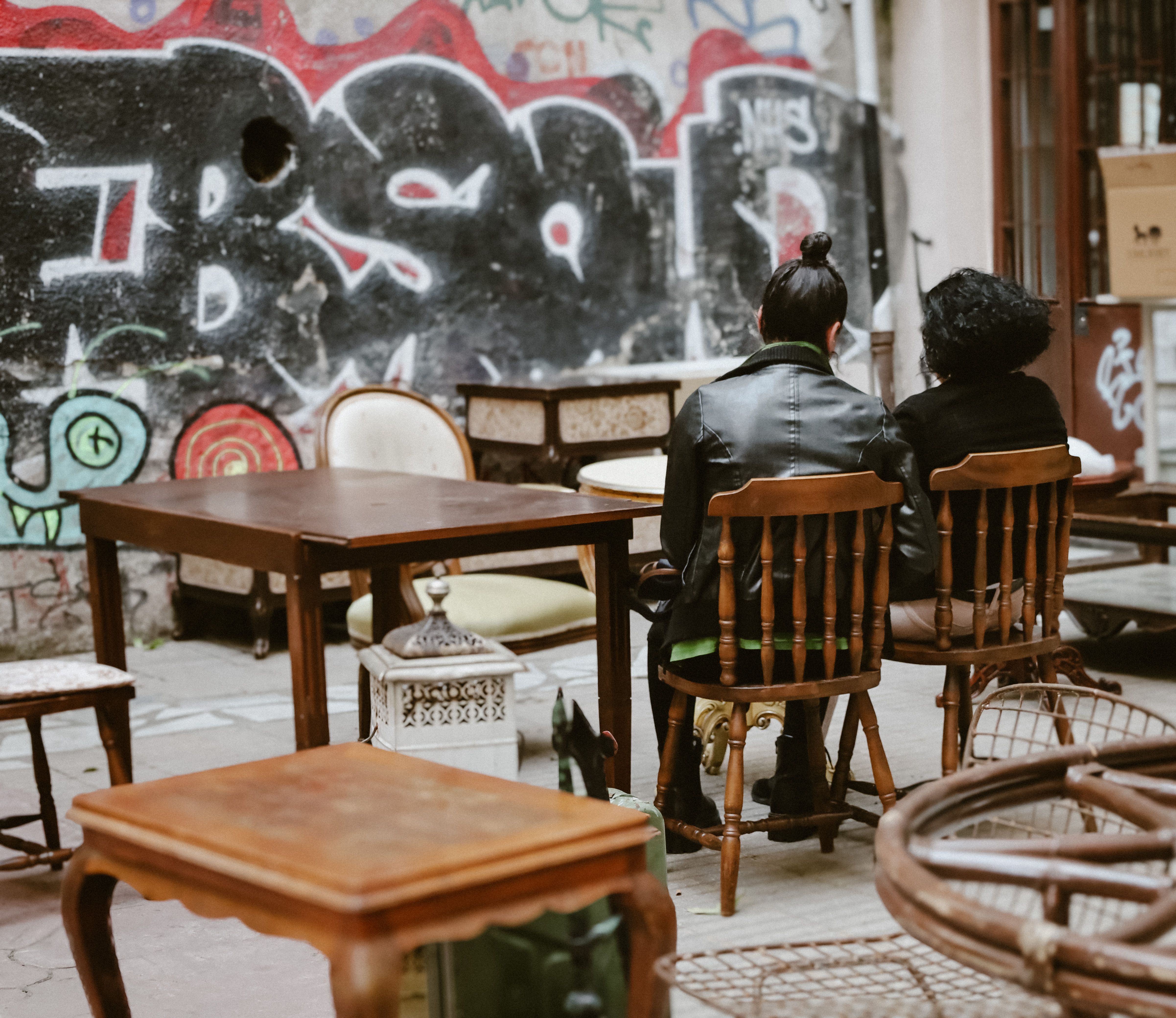Grafiti ve vintage mobilyaların olduğu hareketli bir İstanbul sokağında, ahşap sandalyelerde oturan iki kişi.