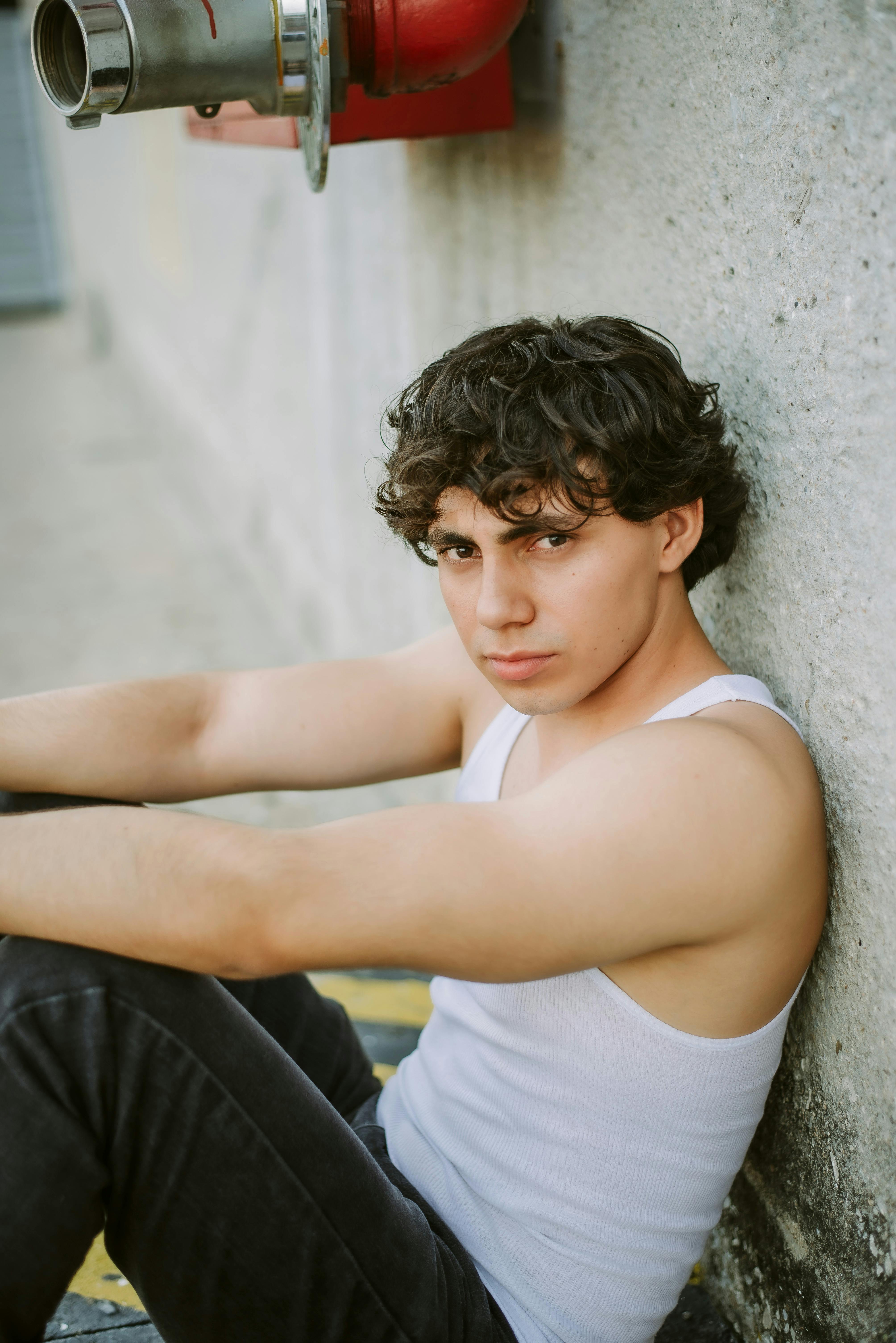 Young man in casual attire sitting thoughtfully against an urban concrete wall.