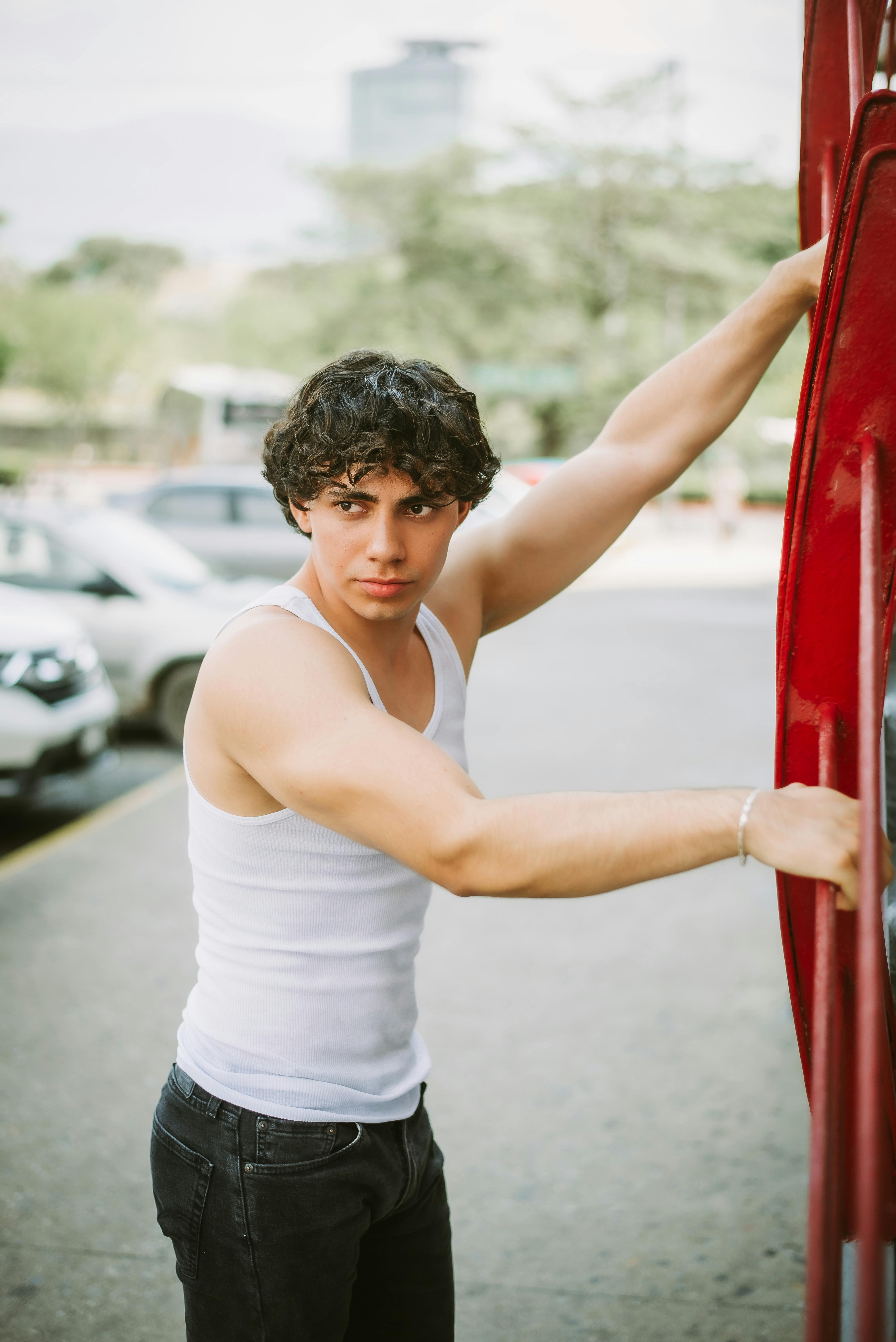 A young man in a white tank top poses outdoors near a red structure.