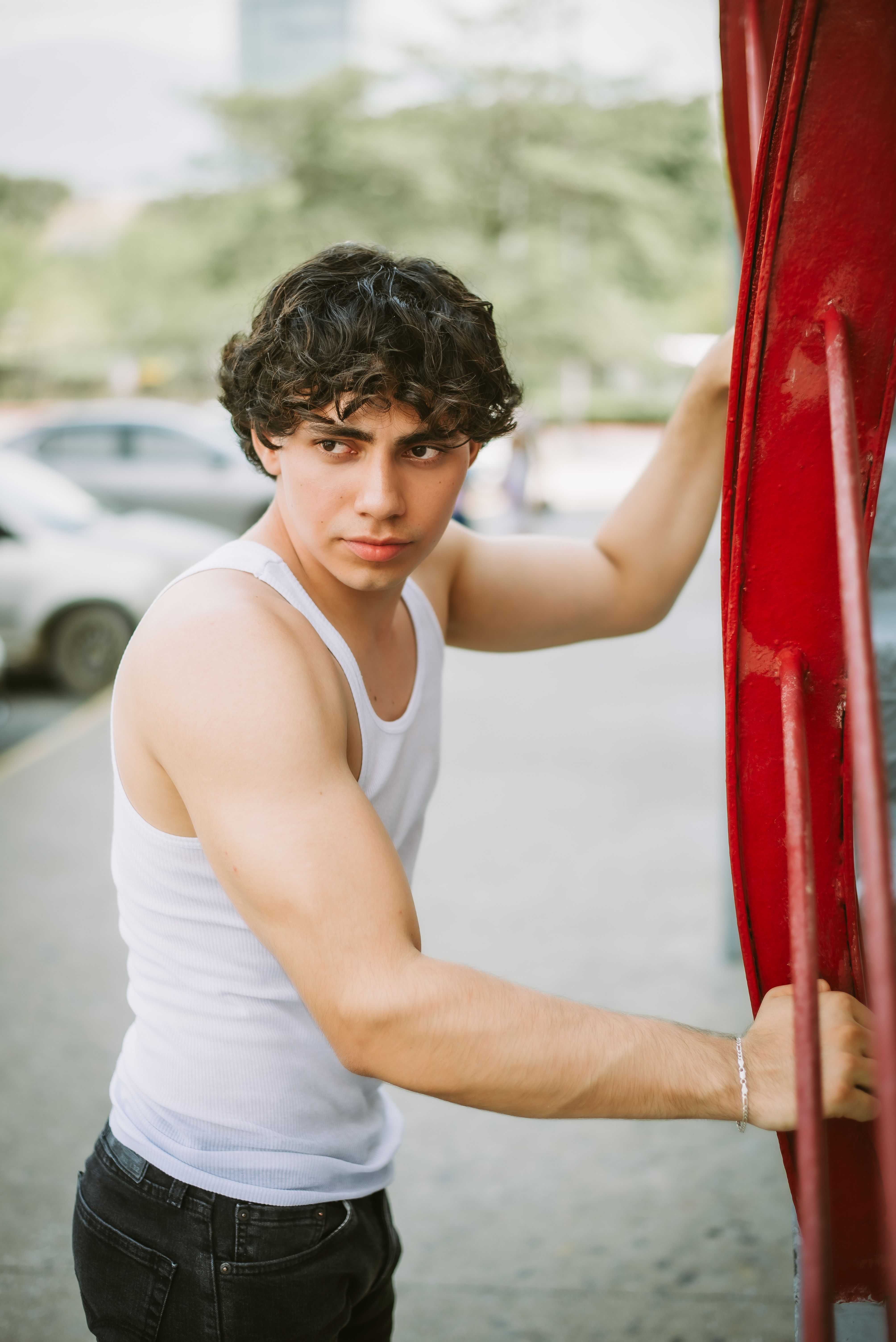 Young man in a white tank top posing with an urban backdrop, exuding confidence.