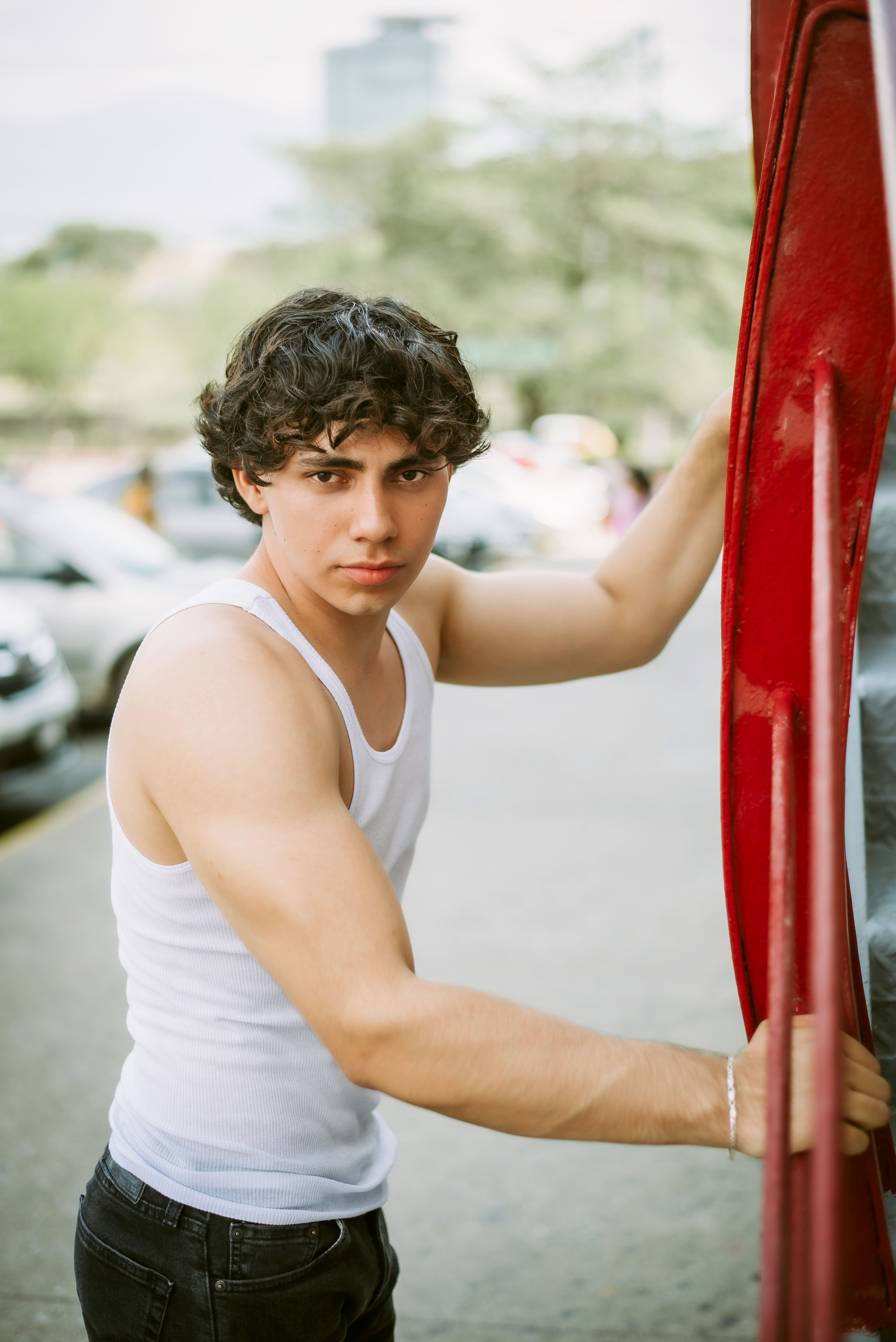 Confident young man in a white tank top exiting a red vehicle on a sunny day.