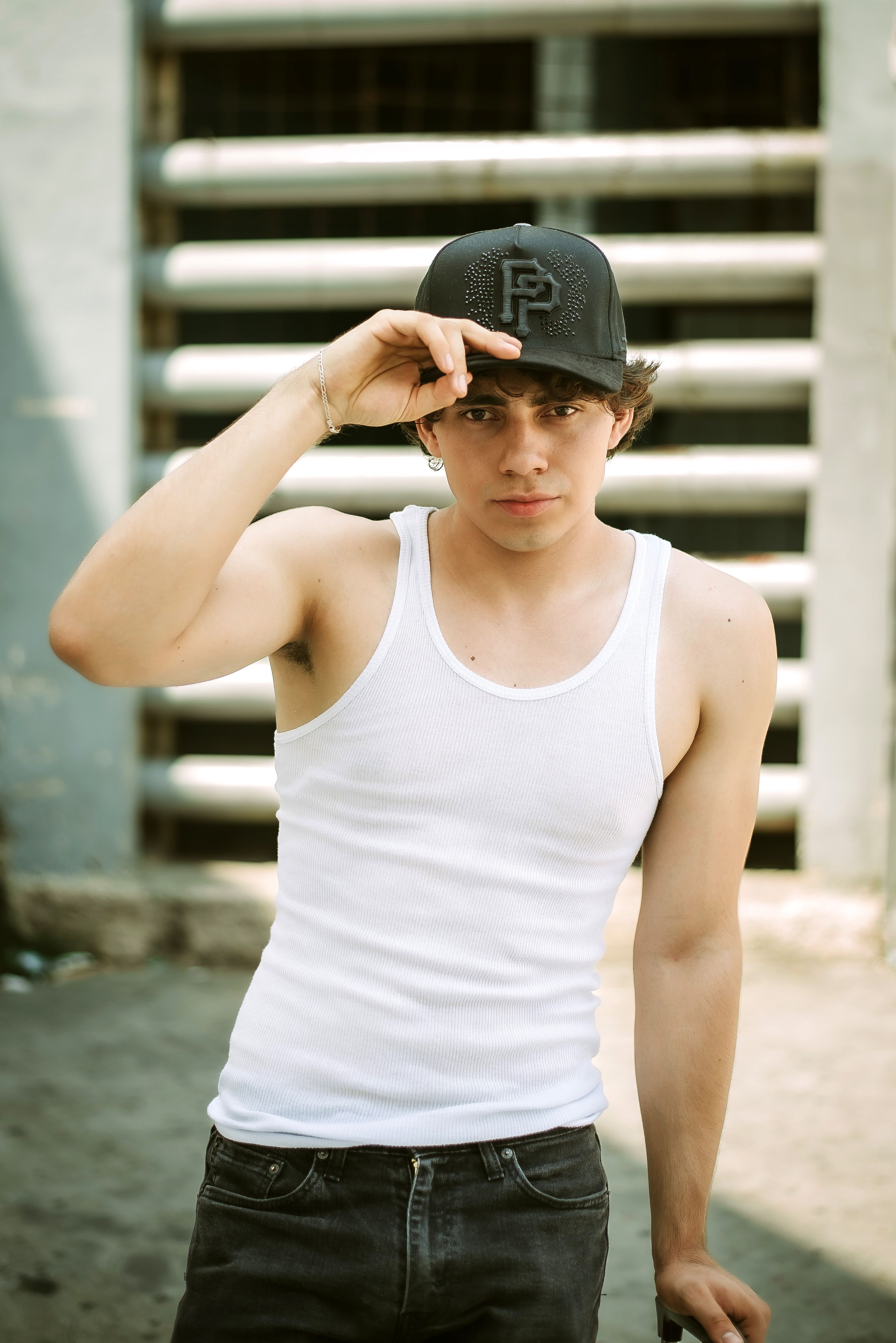 Casual portrait of a young man in a white tank top and black cap, posed in an urban environment.