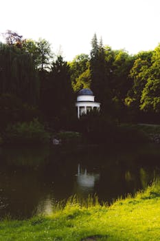 A tranquil scene of a gazebo by a pond surrounded by lush greenery in Calden, Hessen, Germany during sunset.