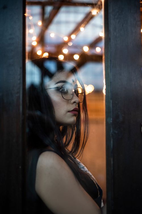 Free A fashionable woman in glasses poses indoors with decorative lights in the background. Stock Photo