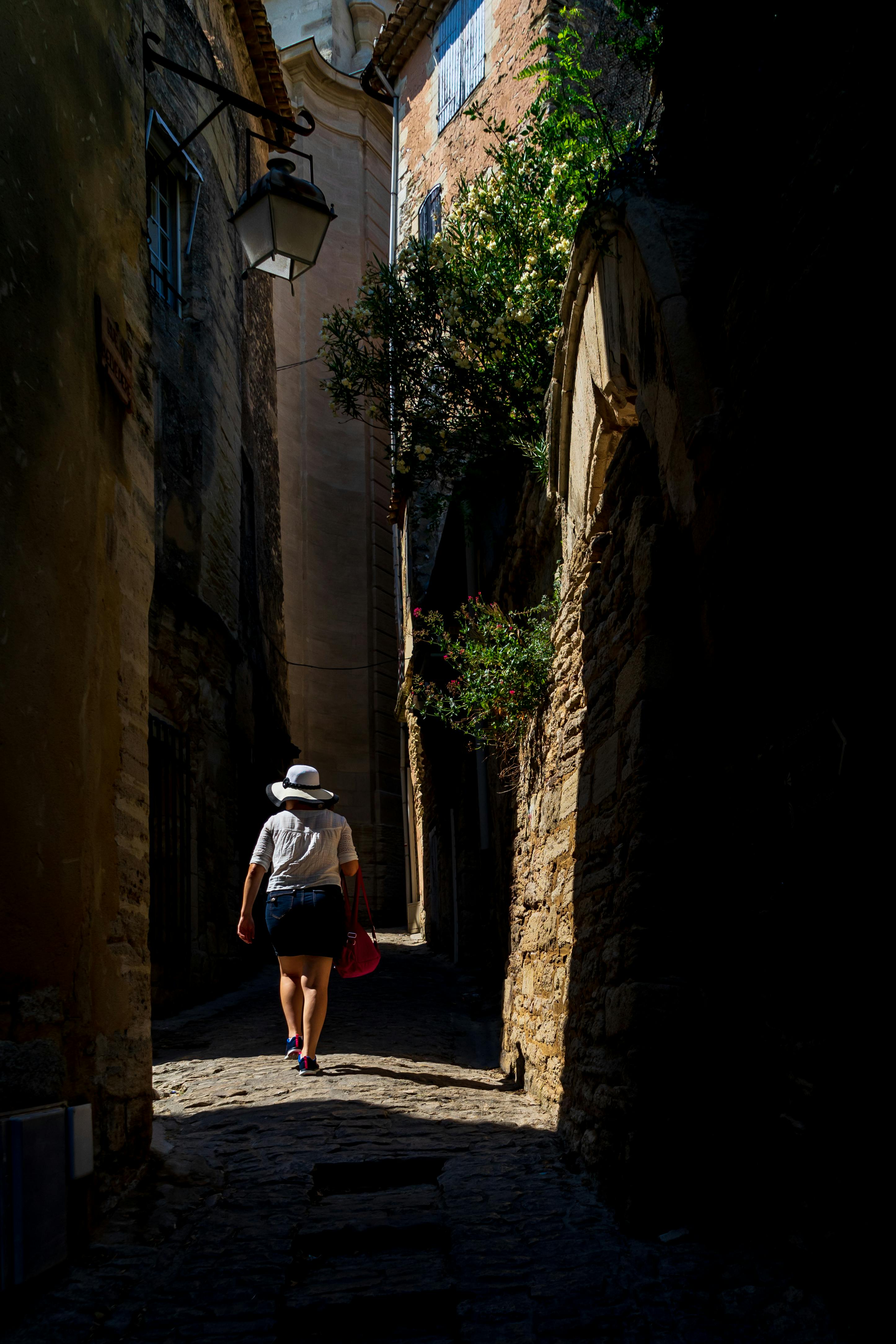 A woman strolls through a narrow, sunlit street in an old European village.