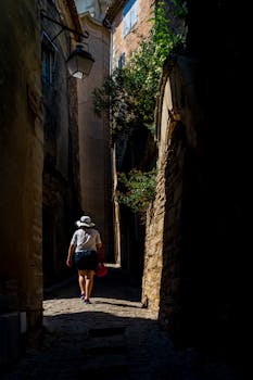 A woman strolls through a narrow, sunlit street in an old European village.