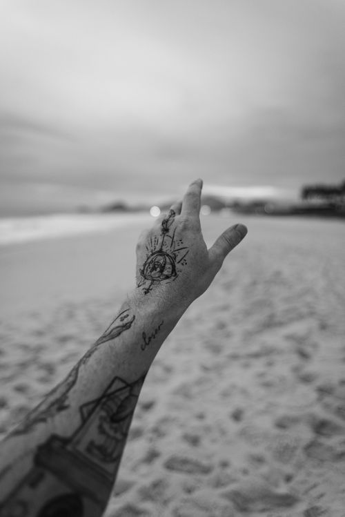 Free A tattooed hand extending towards the beach horizon during twilight, captured in black and white tones. Stock Photo