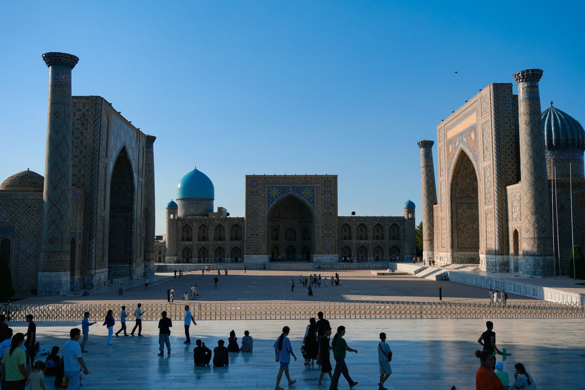 Crowded Registan Square in Samarkand with ancient madrasahs under a clear sky.