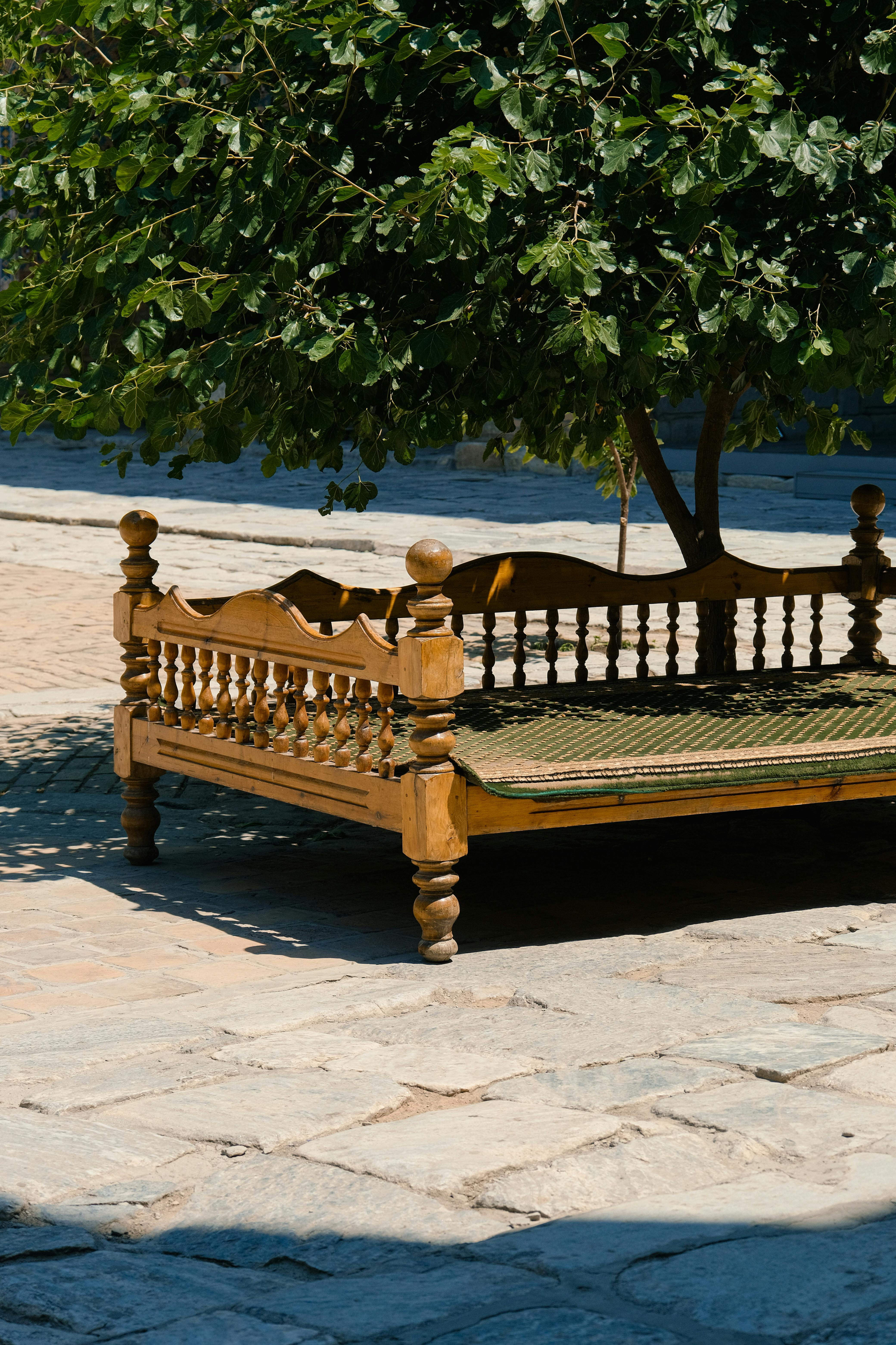 A cozy wooden daybed sits under a lush tree providing shade on a sunny day.
