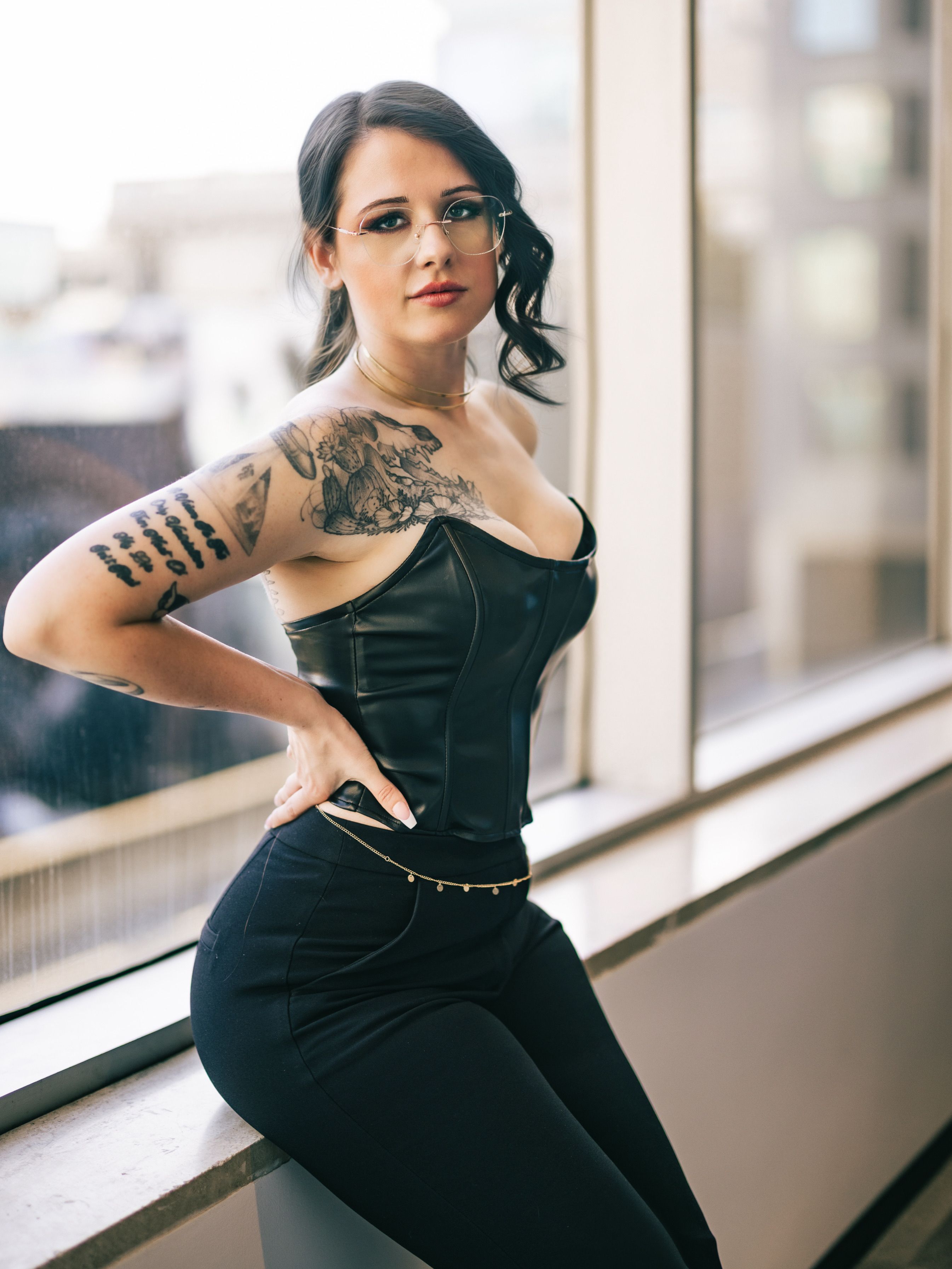 Elegant woman with tattoos in leather corset poses by large window in urban setting.