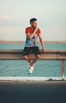 一位身着夏装的时尚男子坐在湖边的栏杆旁，散发着时尚与休闲的气息