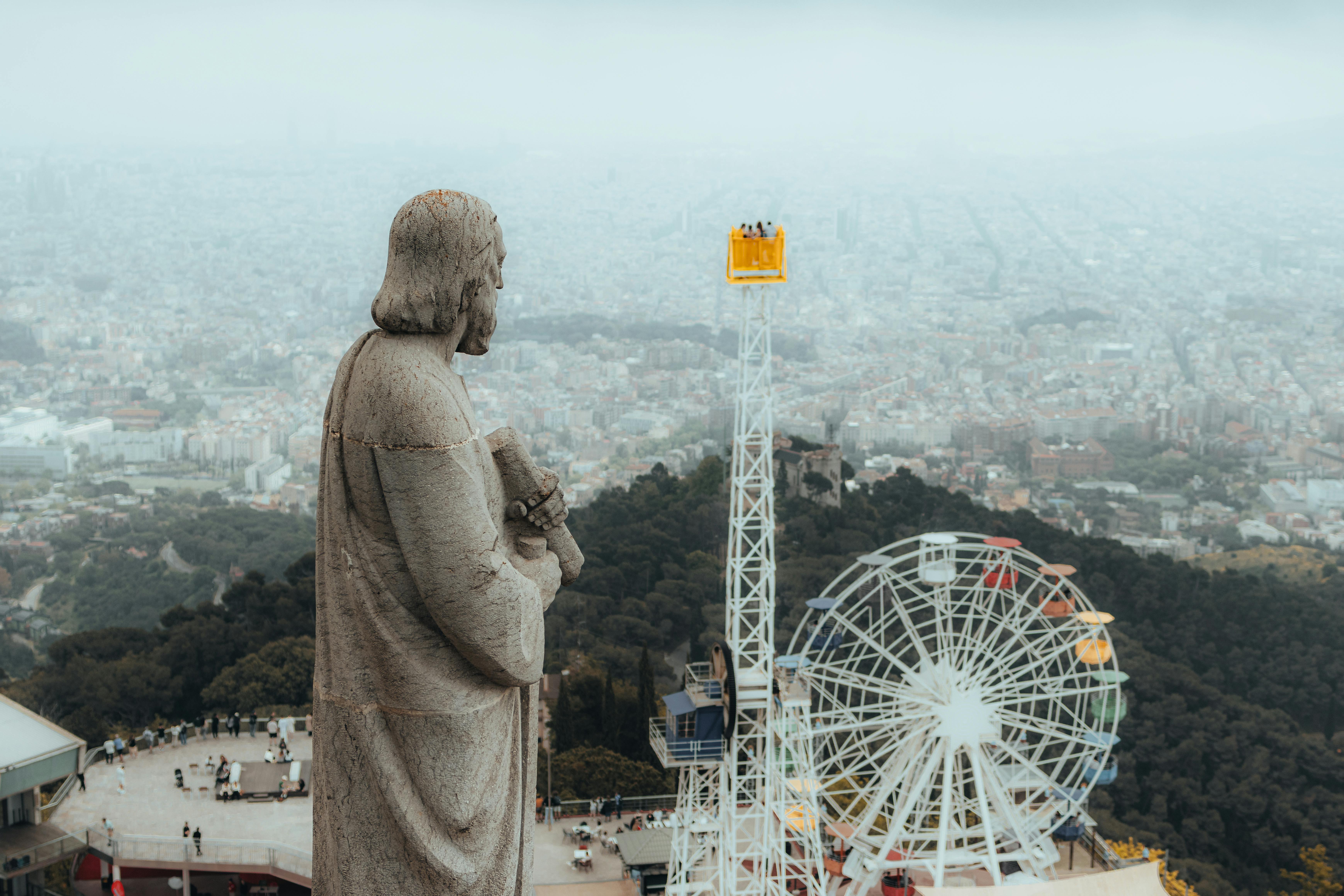 Statue overlooking Barcelona cityscape with Tibidabo Amusement Park and Ferris wheel in foreground.